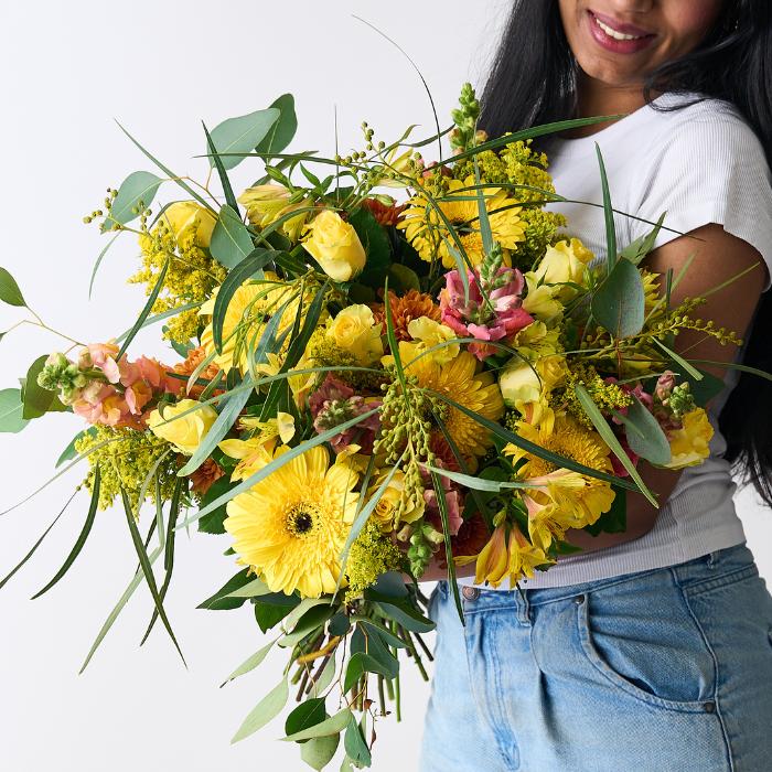 Close-up of a woman holding the Golden Hues Bouquet, showcasing bright yellow gerberas, roses, snapdragons, and fresh greenery.