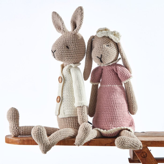 Benedict Bunny Gentleman and Rosie Rabbit Handmade Doll seated together on a wooden bench, showcasing their handcrafted crochet designs in a beige cardigan and pink dress respectively.