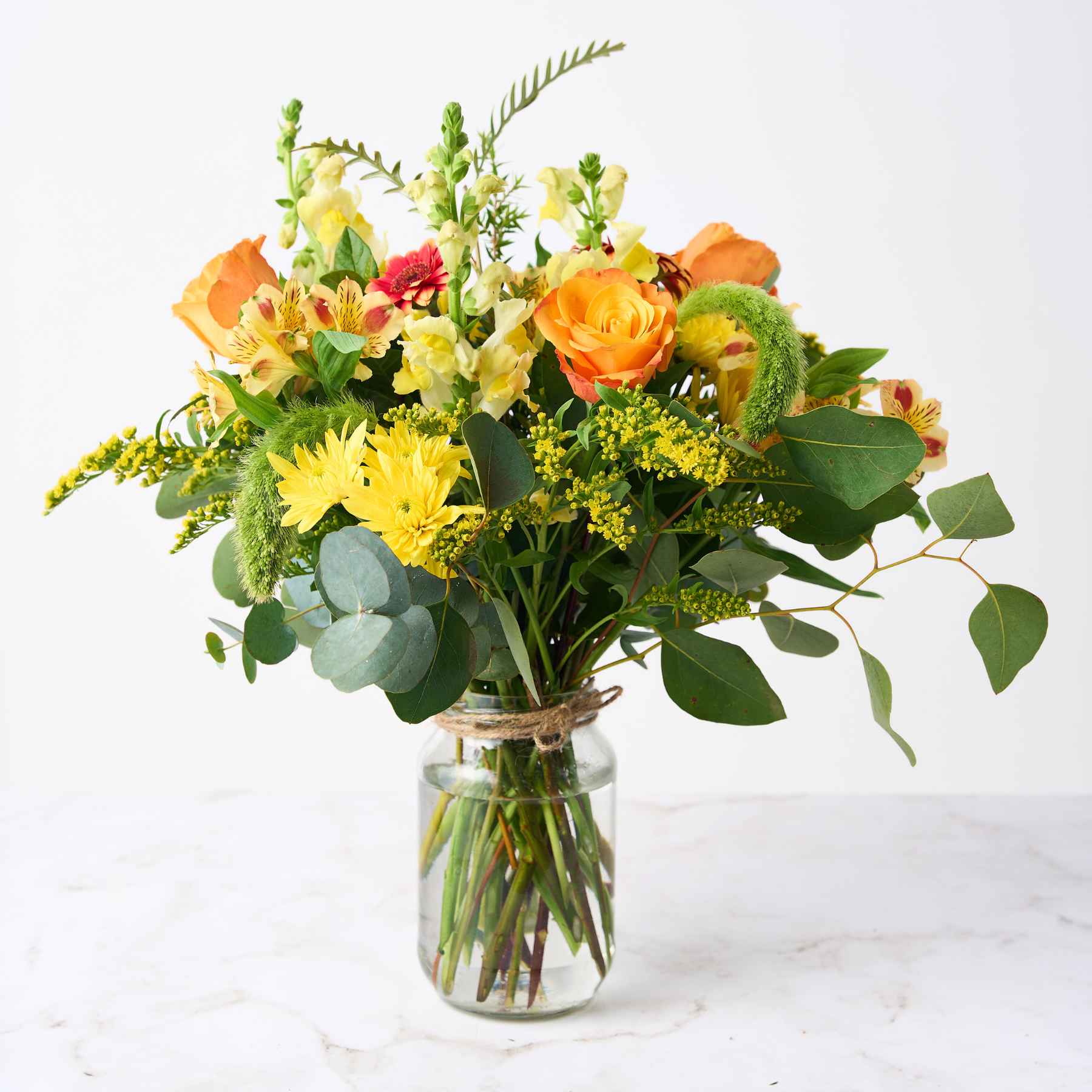 A cheerful flower arrangement in a glass jar, featuring bright yellow chrysanthemums, orange roses, and green foliage, creating a vibrant and happy display.