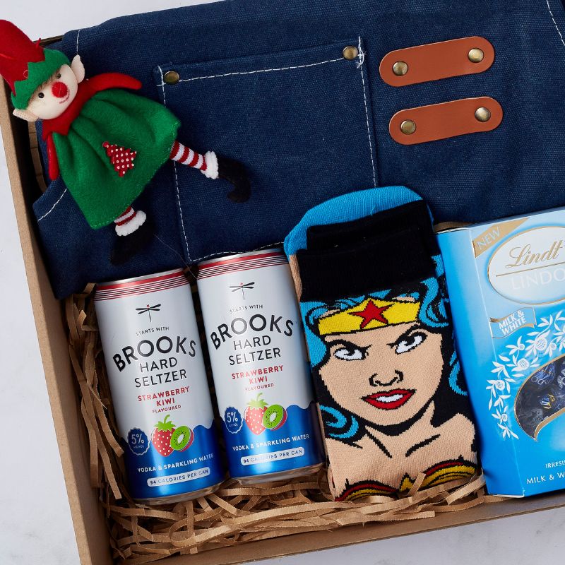 Christmas-themed gift box with Brooks Hard Seltzer, Wonder Woman socks, Lindt chocolates, denim apron, and festive elf plush.