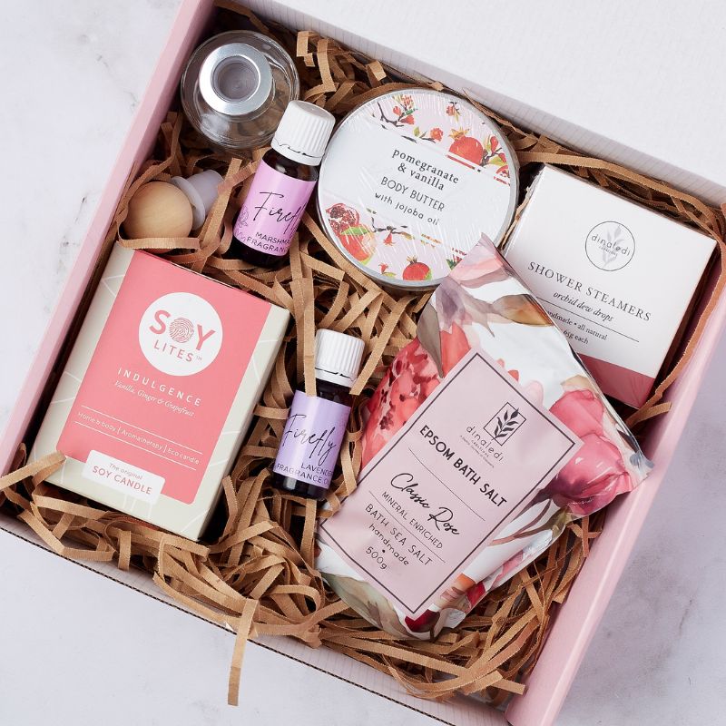 Gift box featuring a selection of self-care products, including a Soy Lites candle, pomegranate and vanilla body butter, Firefly essential oils, Epsom bath salt, shower steamers, and fragrance bottle, all arranged in a pink box with shredded filler.