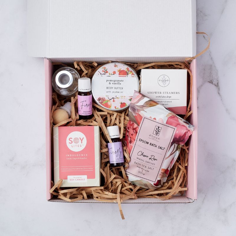 Open gift box showcasing a Soy Lites candle, pomegranate and vanilla body butter, lavender and marshmallow essential oils, shower steamers, cherry rose Epsom bath salt, and a small fragrance bottle, arranged neatly on shredded paper filler.