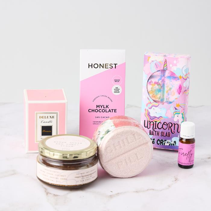 A beautifully arranged collection of self-care products on a white surface. The items include a pink box of Honest Mylk Chocolate, a pink and gold jar of brown sugar coffee scrub, a pink deluxe vanilla-scented candle, a round pink bath bomb, a colourful Unicorn Bath Slab, and a small bottle of Firefly coconut fragrance oil. The packaging is predominantly pink with playful and vibrant designs, creating a fun, indulgent, and pampering vibe.