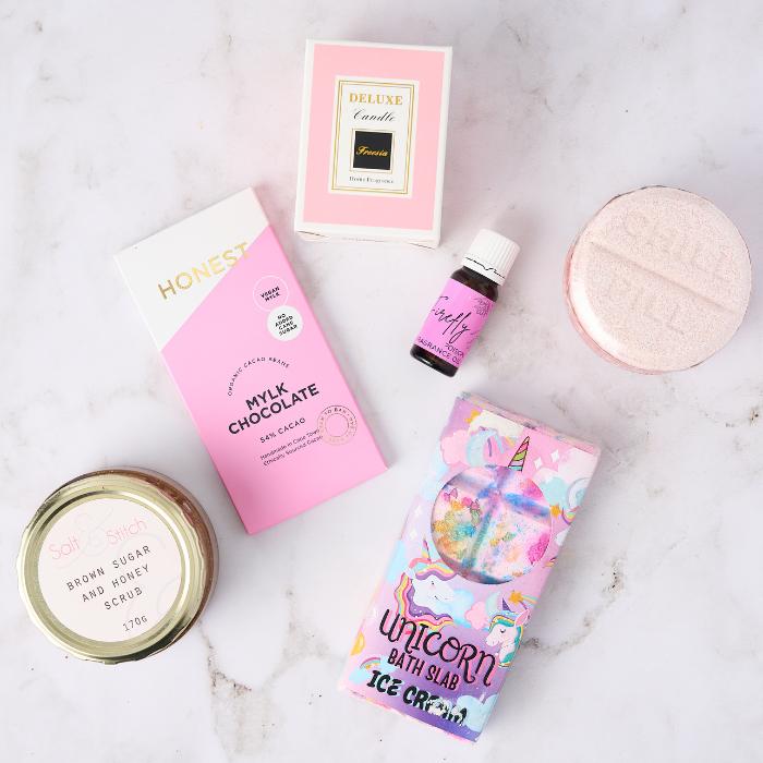 A flat lay of luxurious self-care products displayed on a white marble surface. The collection features Honest Mylk Chocolate in pink packaging, a deluxe vanilla-scented candle in a soft pink box, Firefly coconut fragrance oil, a colourful Unicorn Bath Slab, a pink round bath bomb, and a jar of brown sugar and honey scrub. The arrangement evokes a feeling of pampering and indulgence, with vibrant and playful packaging elements.