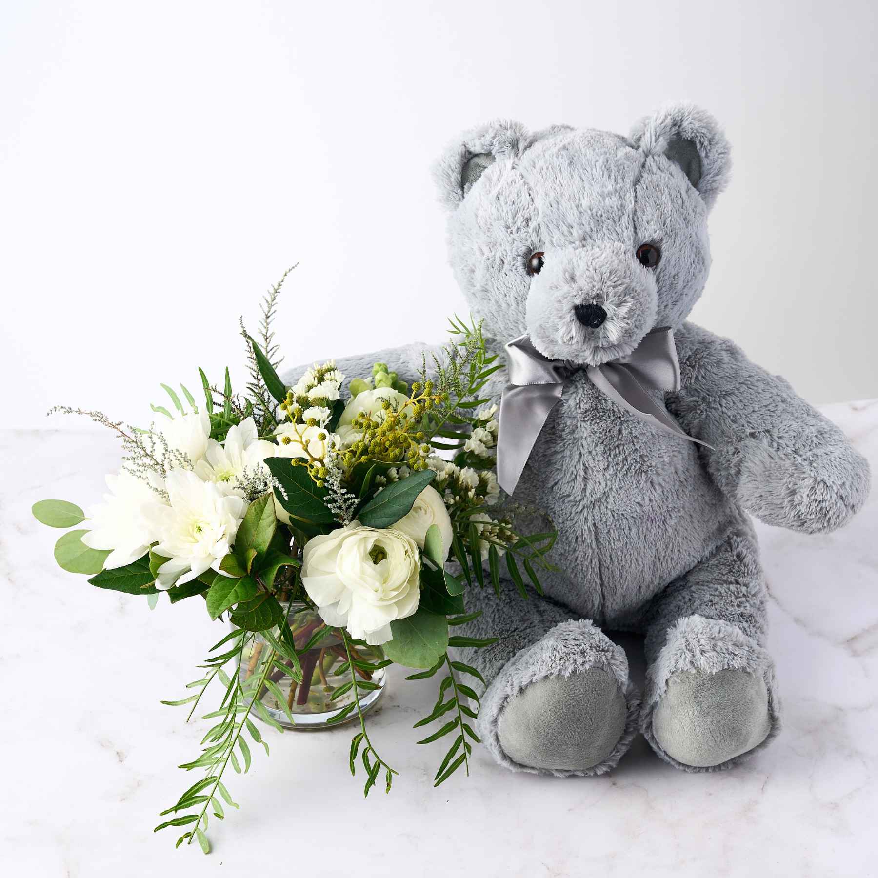 A soft grey teddy bear with a silver ribbon, sitting beside a bouquet of white flowers and greenery, representing the 'It's a Boy' gift arrangement.