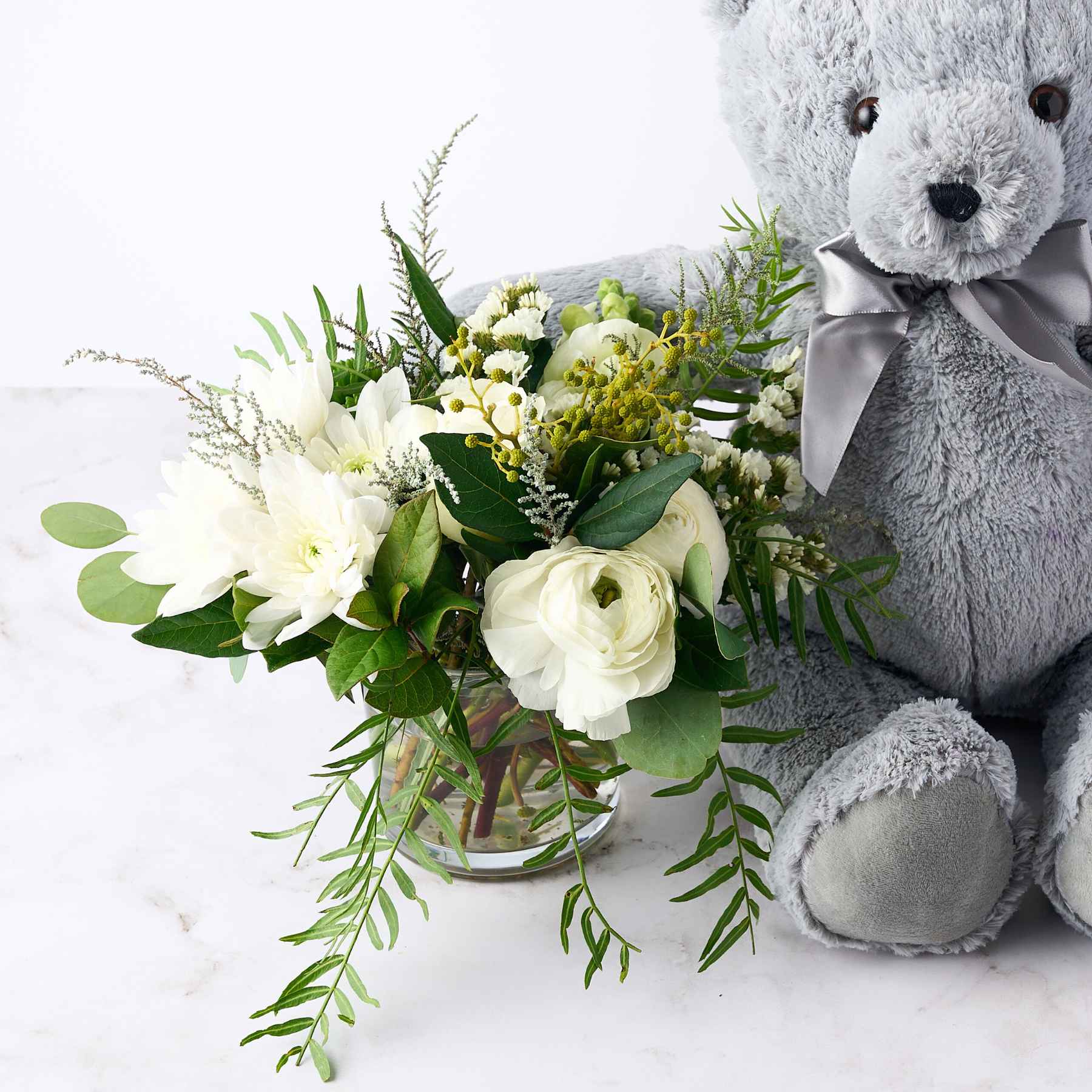 Close-up of a grey teddy bear with a bouquet of white flowers and green foliage, symbolising the celebration of a baby boy with the 'It's a Boy' gift.