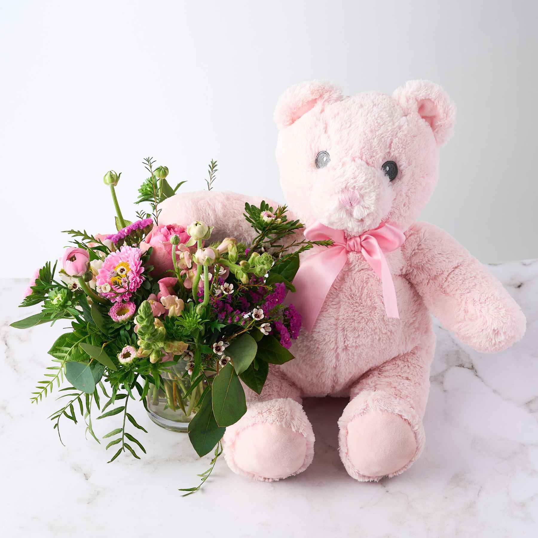 A soft pink teddy bear with a ribbon, sitting beside a bouquet of pink flowers and greenery, representing the 'It's a Girl' gift arrangement.