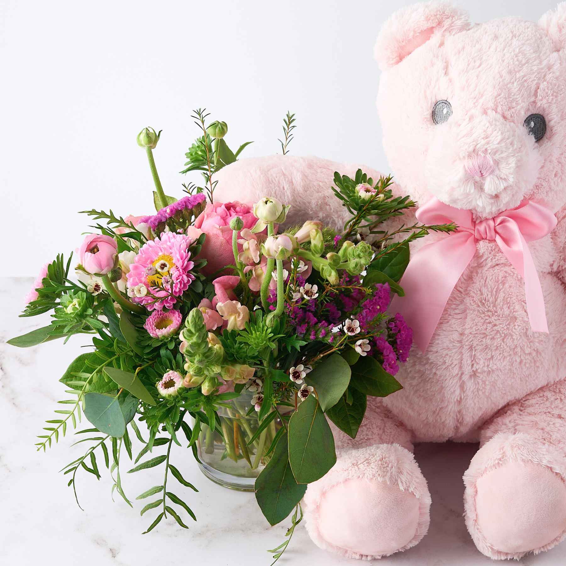 Close-up of a pink teddy bear with a bouquet of pink and green flowers, symbolising the celebration of a baby girl with the 'It's a Girl' gift.