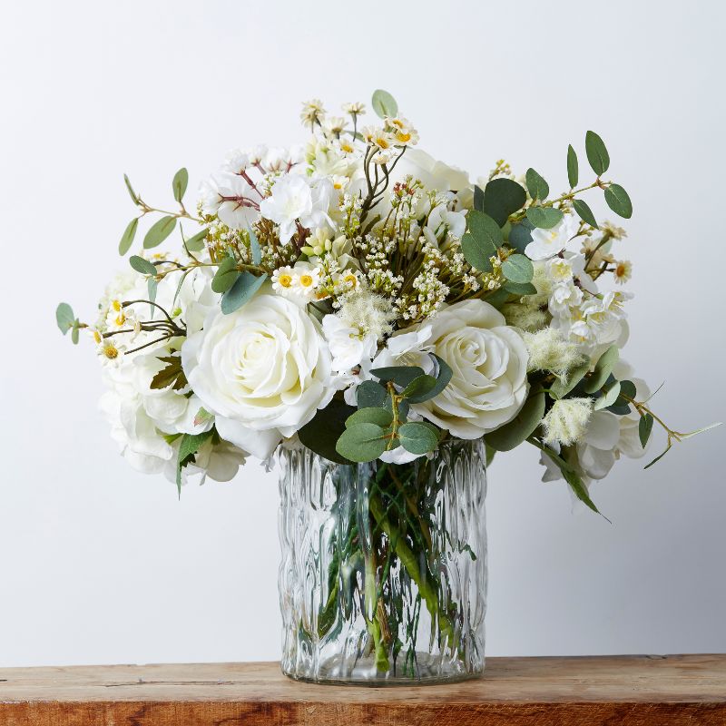 Elegant ivory artificial floral arrangement in a textured glass vase, featuring white roses, daisies, eucalyptus, and mixed greenery.