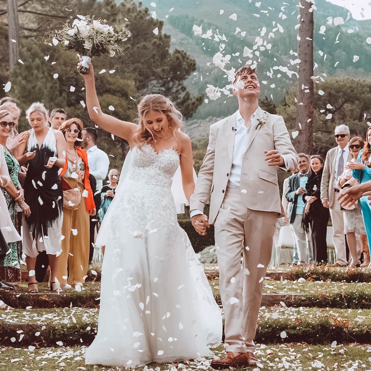 Bride Chloe and groom James walk down the aisle outdoors, smiling and holding hands, with guests showering them with flower petals. Weddings. Fabulous Flowers and Gifts.