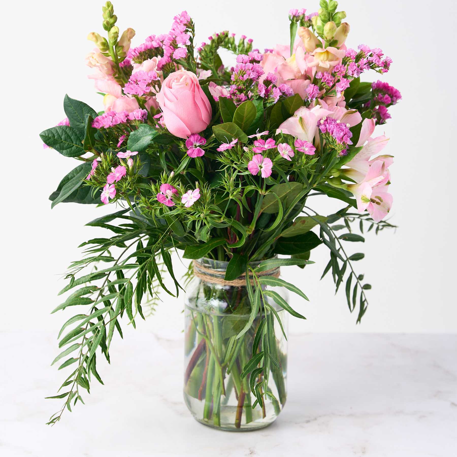 A charming flower arrangement in a glass jar, featuring pink roses, snapdragons, and various greenery, creating a delightful and cheerful display.