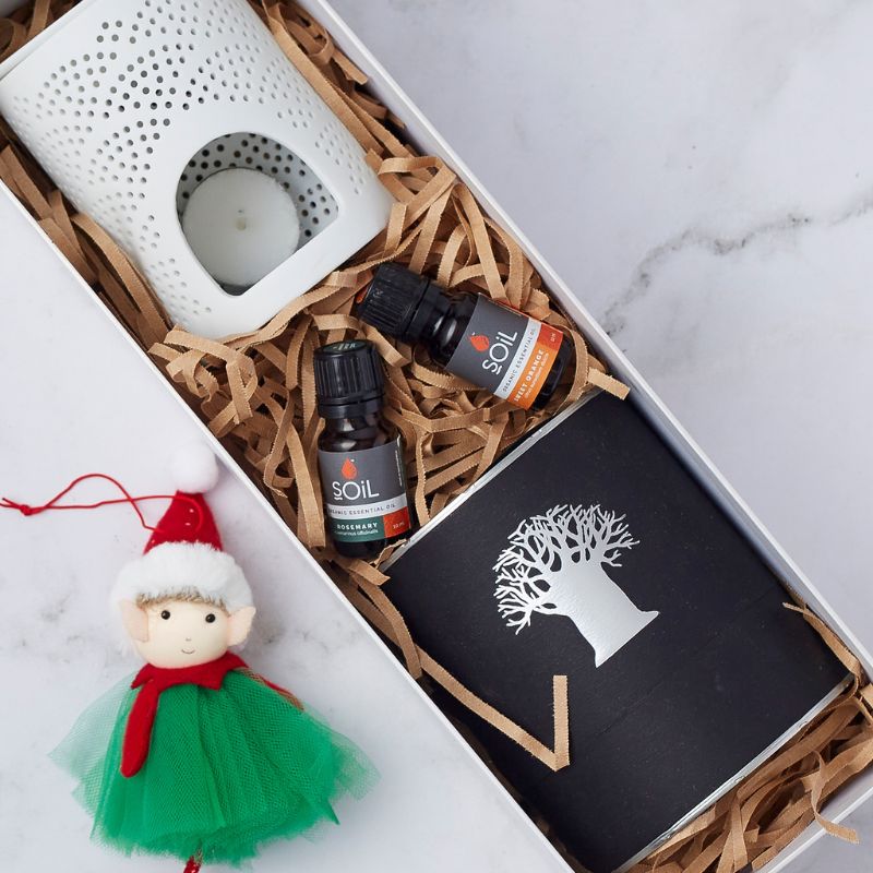 Festive gift box featuring a ceramic candle holder, two Soil essential oils (rosemary and orange), a black candle with a silver tree design, and a small elf ornament dressed in a green tulle skirt.