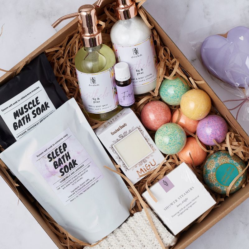 Close-up of a pamper gift box with bath soaks, body wash, bath bombs, shower steamers, and a heart-shaped lollipop.