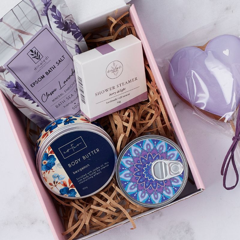 Close-up of luxury gift box featuring lavender bath salts, berry shower steamer, bergamot body butter, candle, and heart cookie.