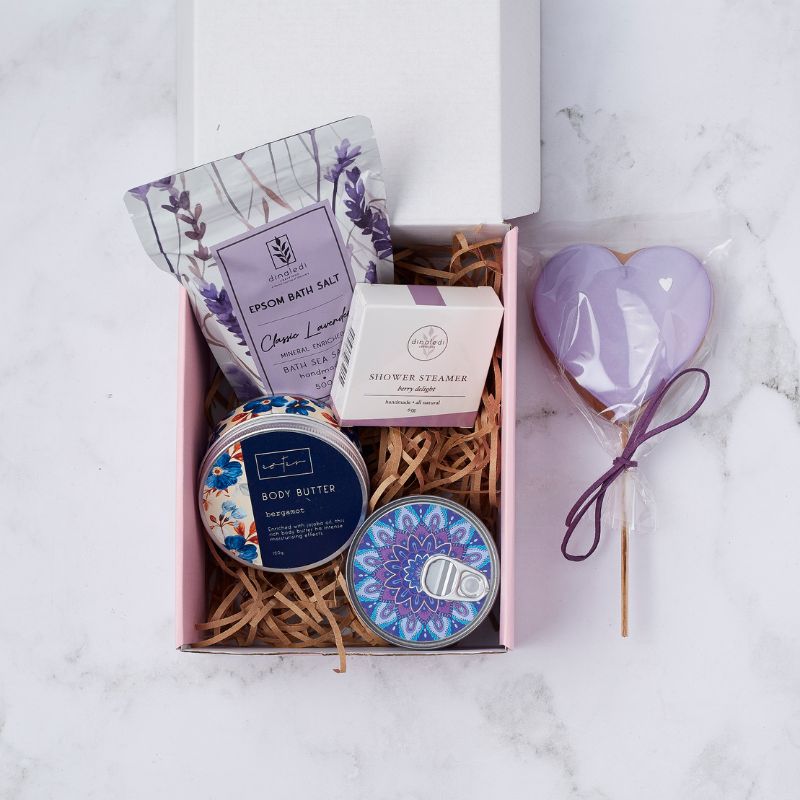 Gift box with lavender-scented bath salts, berry shower steamer, bergamot body butter, decorative candle, and heart-shaped purple cookie.