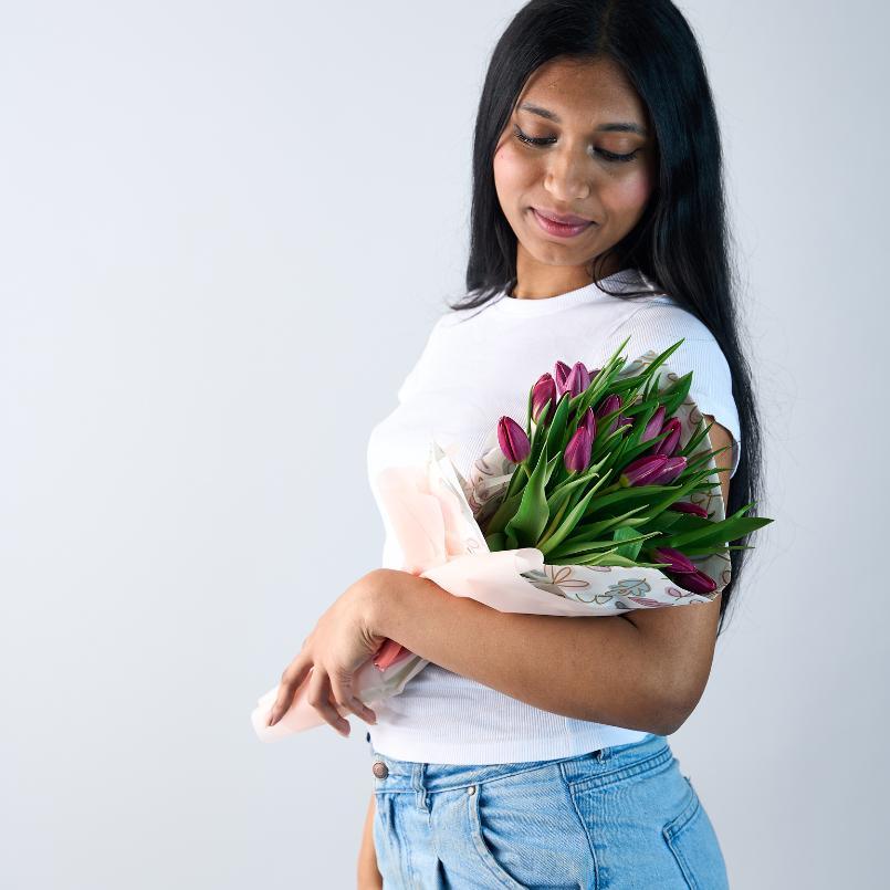 Woman holding a bouquet of purple tulips wrapped in soft pastel floral paper, wearing a white t-shirt and jeans.