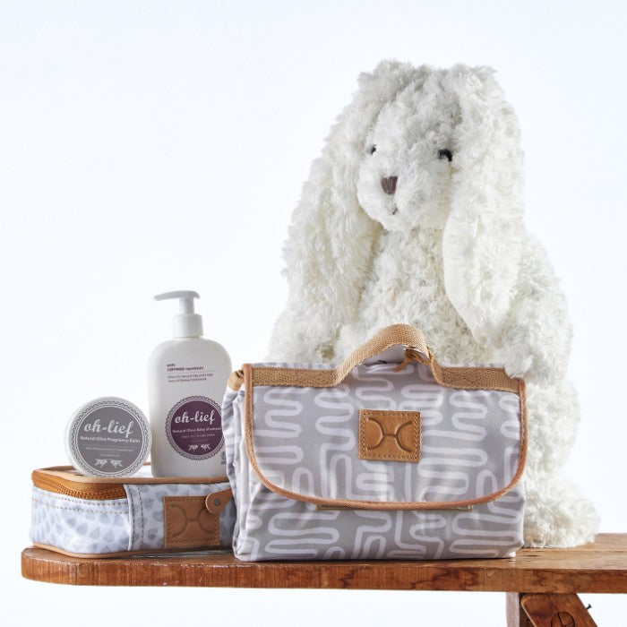 Little Luxuries Baby Gift Set featuring a fluffy white bunny plush, organic baby skincare products, and a stylish grey patterned diaper bag.