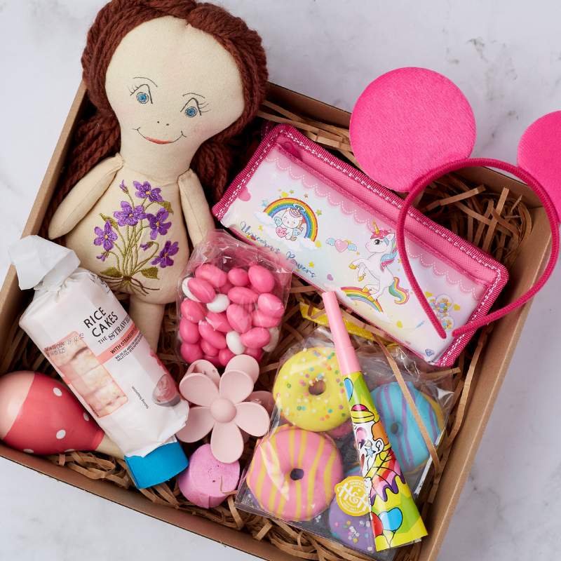 Close-up of the Little Princess Gift Box with a doll, pink mouse-ear headband, unicorn pencil case, rice cakes, and fun pink and yellow treats.