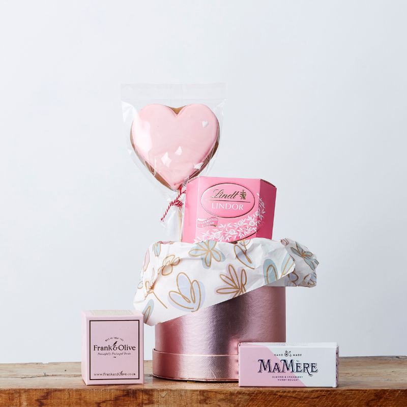 A beautifully arranged gift box featuring a pink heart-shaped biscuit lollipop, Lindt Swiss milk chocolate truffles in a pink box, a Frank & Olive treat box, and a Ma Mère almond & cranberry nougat bar, all elegantly displayed in a decorative round box with floral-patterned tissue paper.