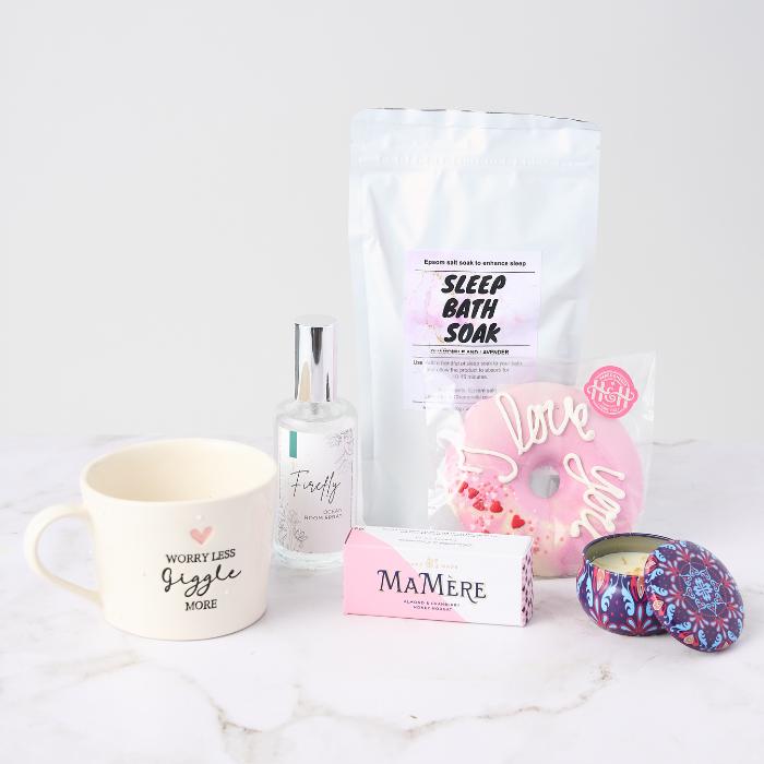 Lovely Lady Gift Set featuring a 'Worry Less, Giggle More' mug, sleep bath soak, room spray, donut-shaped bath bomb, nougat treat, and a decorative candle tin.