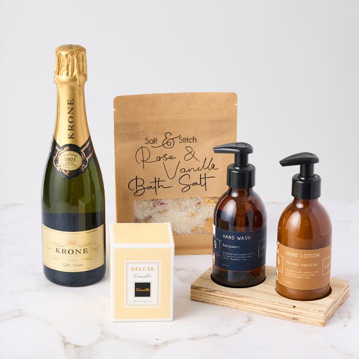 A luxurious pamper gift set displayed on a white marble surface. The set includes a bottle of Krone Vintage Rosé Sparkling Wine, Salt & Stitch Rose & Vanilla Bath Salt in a kraft pouch, a deluxe vanilla-scented candle in a cream box, and matching bergamot-scented hand wash and velvet vanilla hand lotion in amber bottles with black pump tops, sitting on a wooden tray.