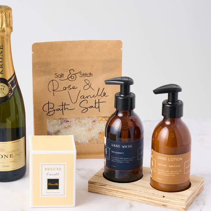 Close-up of the Luxe Living pamper gift set featuring a bottle of Krone Vintage Rosé Sparkling Wine, Salt & Stitch Rose & Vanilla Bath Salt in a kraft pouch, a deluxe vanilla-scented candle in a cream box, and a hand care duo of bergamot hand wash and velvet vanilla hand lotion in amber bottles with pump dispensers on a wooden stand.