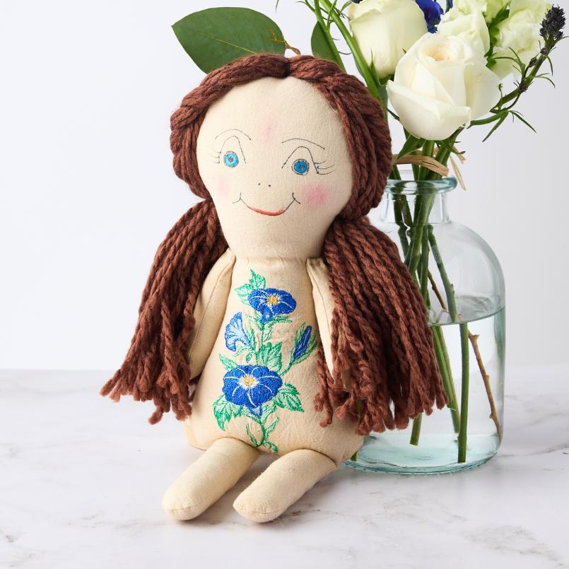 Handcrafted Morning Glory Dreams Doll with blue morning glory embroidery, seated next to a vase of white roses and blue blooms.