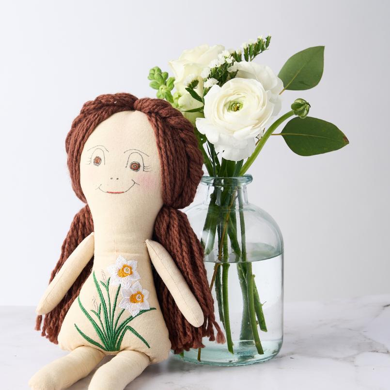 Close-up of the New Beginnings Gift Set, showcasing a handcrafted doll with floral embroidery and a vase of white roses and ranunculus flowers.