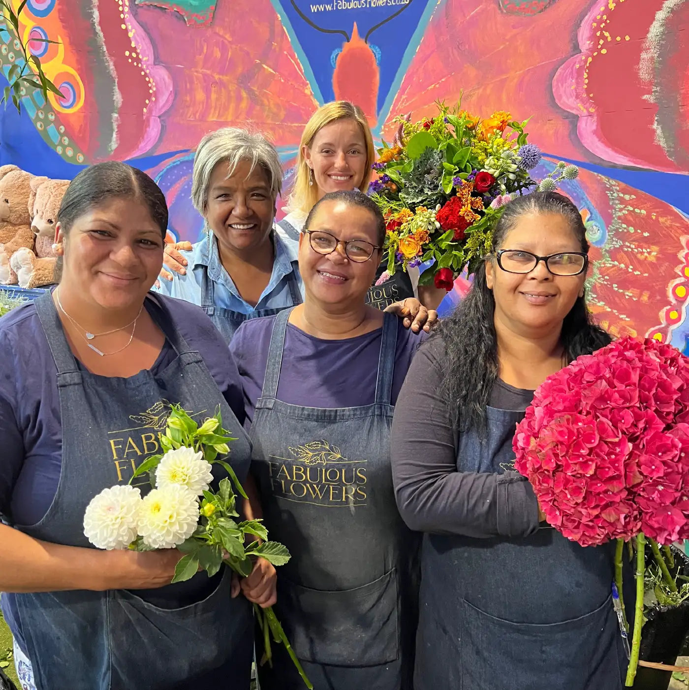 Team of smiling florists holding vibrant flower arrangements, standing in front of a colourful mural. Text highlights Experience Fabulous Flowers, luxury meets responsibility, and the art of floristry. Our Difference. Fabulous Flowers and Gifts.