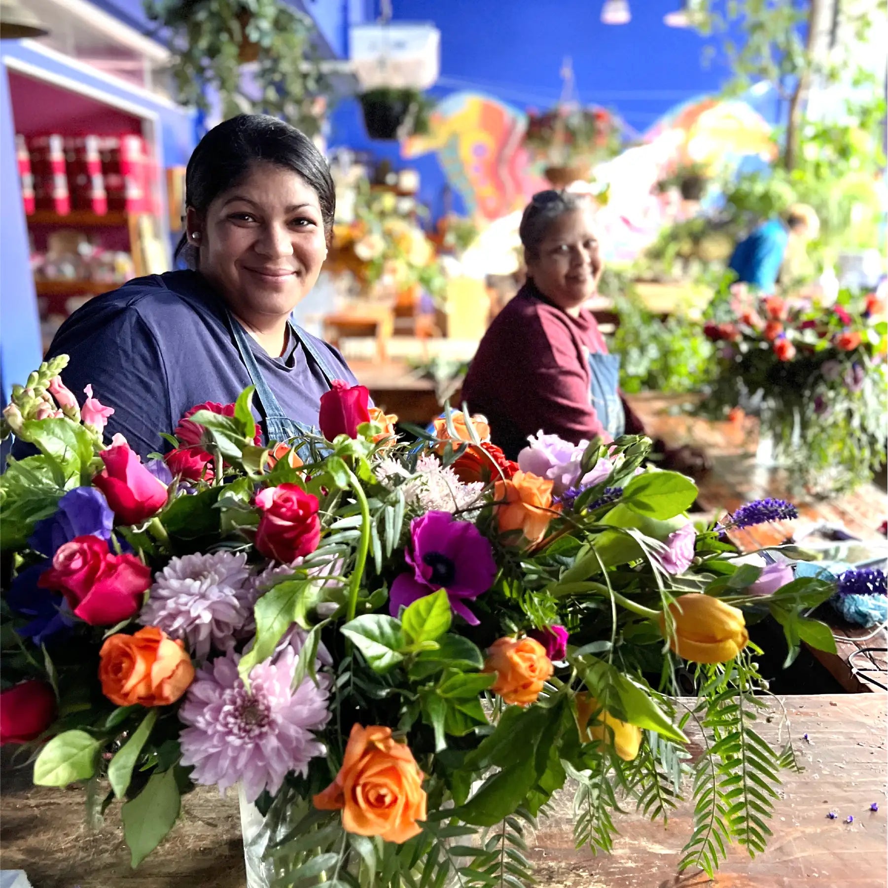 Smiling florist arranging a colourful bouquet of mixed flowers, with another florist working in the background. Text highlights Embracing Kindness, community service, and employee engagement. Our Difference. Fabulous Flowers and Gifts.