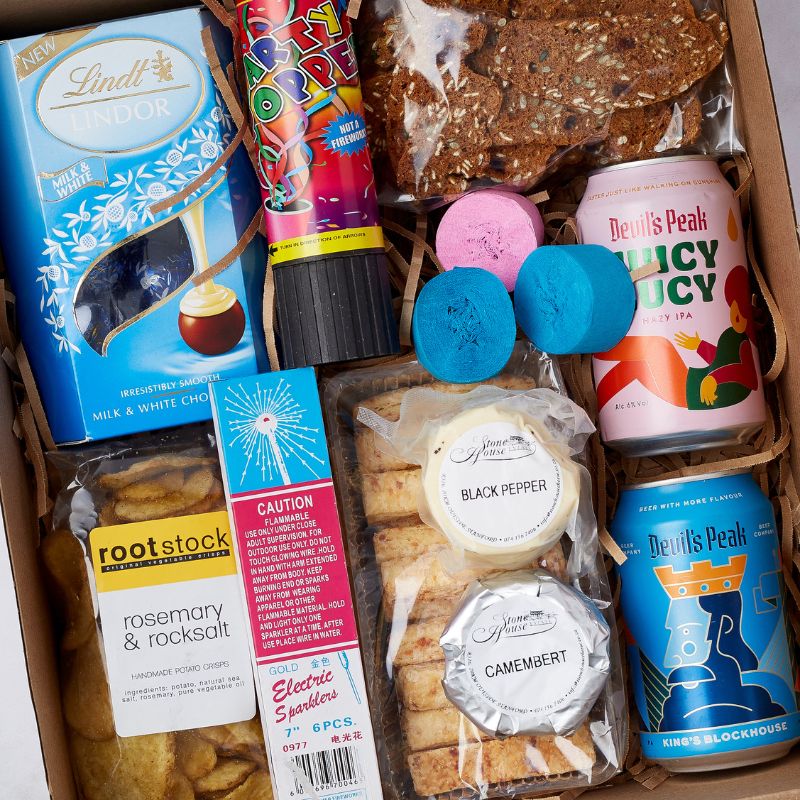 Close-up of a party gift box featuring Lindt chocolates, Devil's Peak craft beers, cheese crackers, rootstock crisps, sparklers, and colourful party decorations.