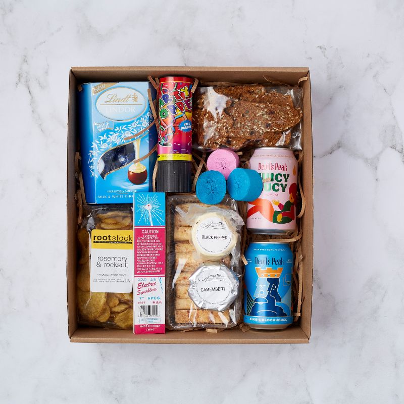 Top view of a festive party gift box with Lindt chocolates, Devil's Peak craft beers, cheese crackers, rootstock crisps, sparklers, and party poppers.