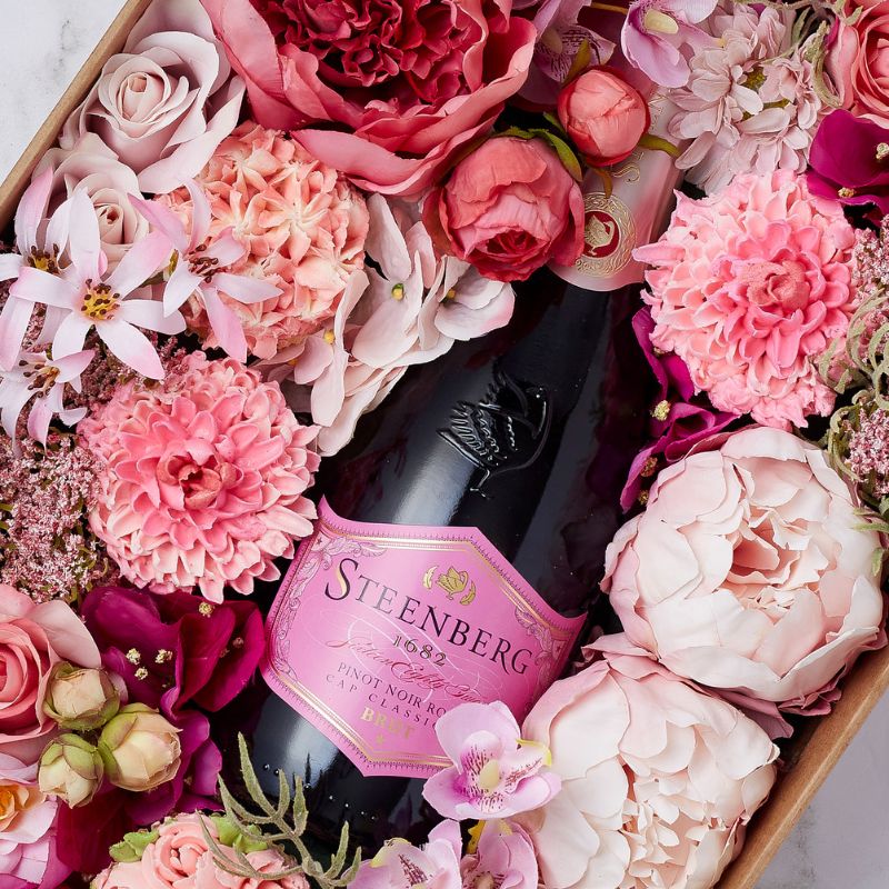 Close-up of a Steenberg Pinot Noir Brut Rosé bottle surrounded by vibrant pink and blush flowers in a luxury gift box.