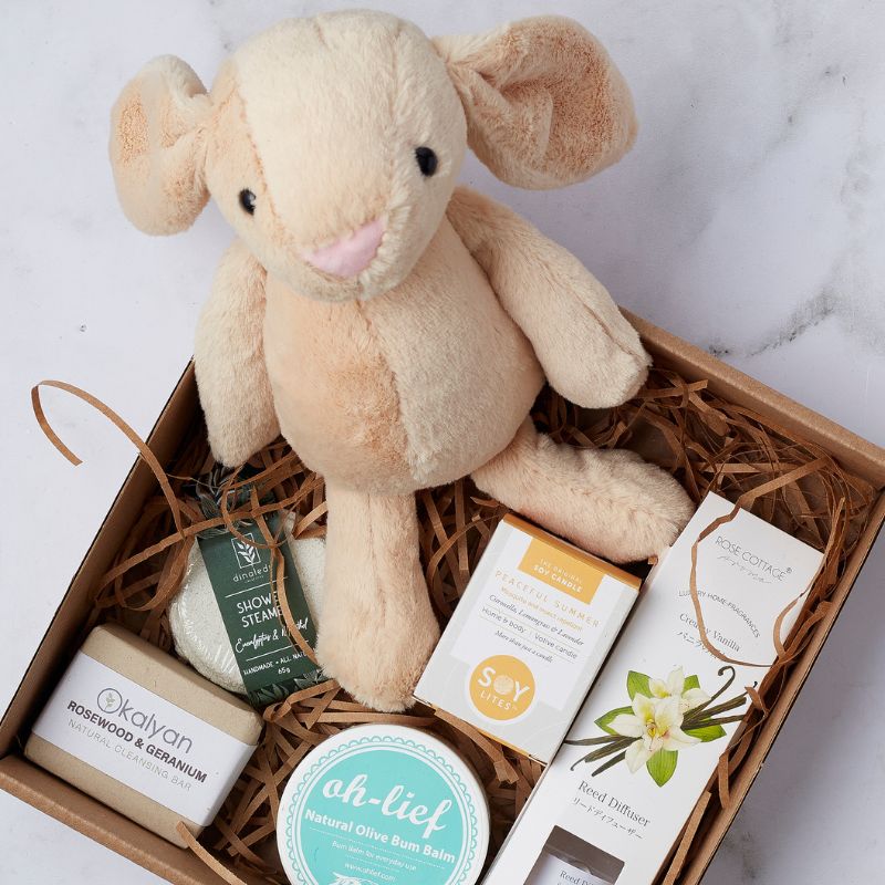 Close-up of gift box featuring plush toy, cleansing bar, soy candle, reed diffuser, and natural olive bum balm