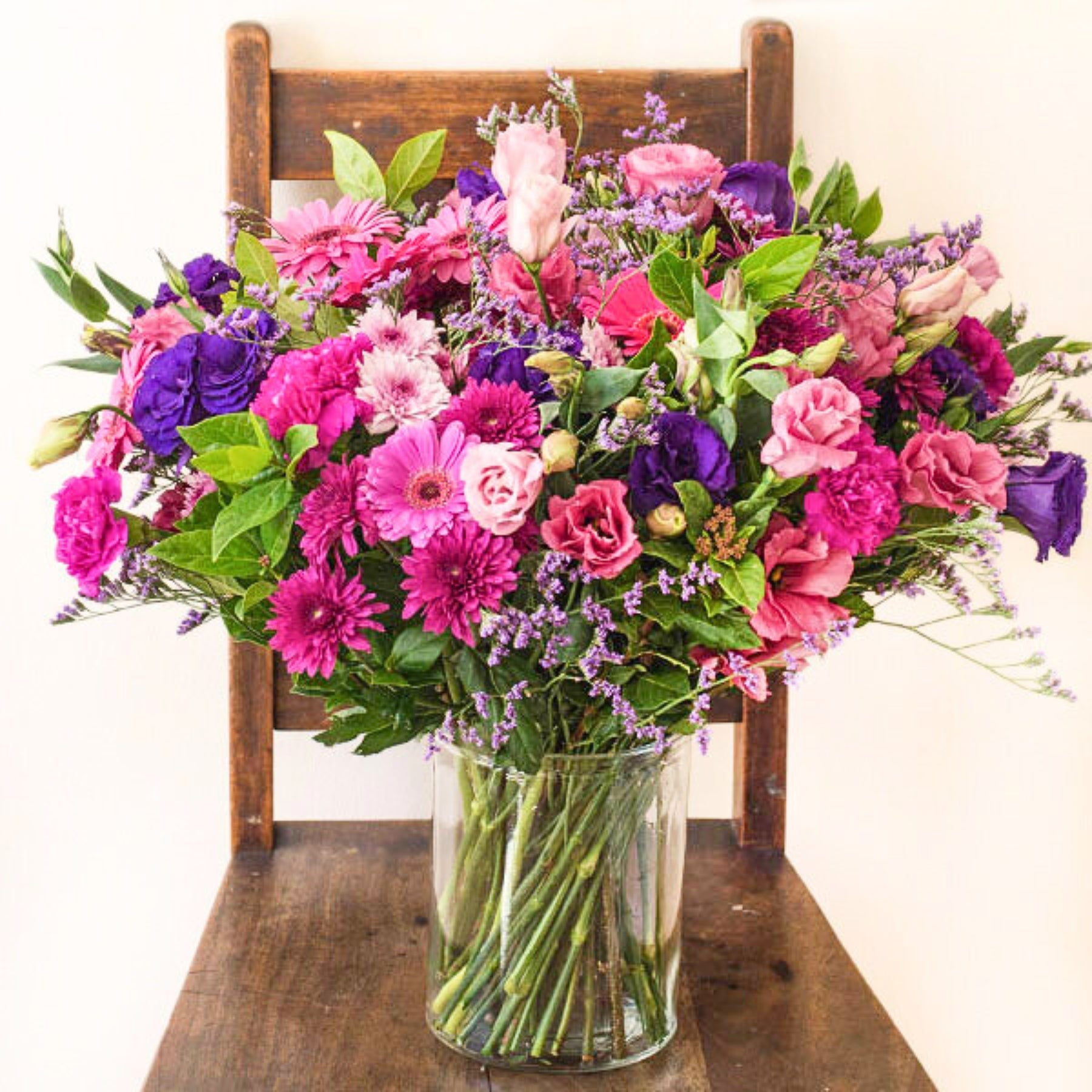 Lush pink and purple flower arrangement in clear vase, showcased on a rustic wooden chair.