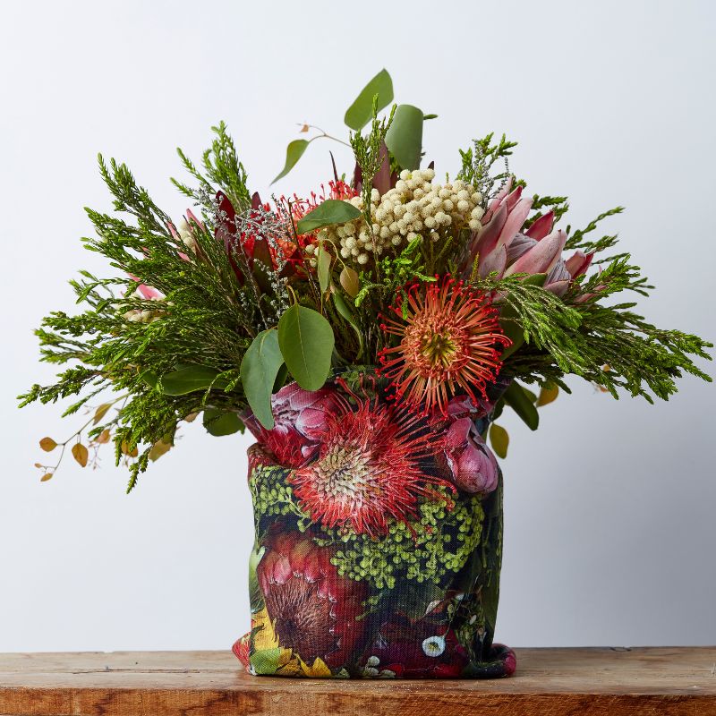 Fresh floral arrangement featuring South African proteas, pincushions, and lush greenery in a protea-patterned fabric wrap.