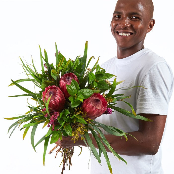 Protea Garden Flower Bouquet held by a smiling man, featuring rich red proteas and lush greenery.