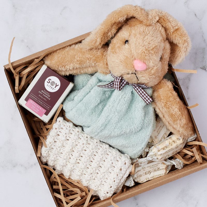 Close-up of a gift box containing a plush bunny, crocheted blanket, Soy Lites candle, and macadamia nougats.