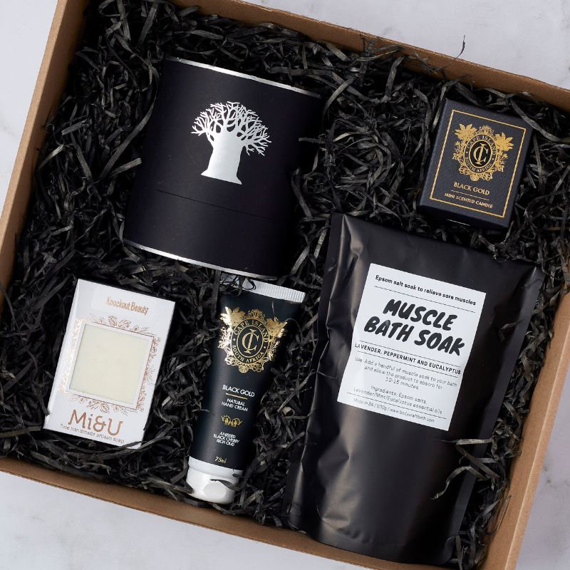 Close-up of Recharge Pamper Gift Set contents, including bath soak, hand cream, candle, and handcrafted soap in black packaging.