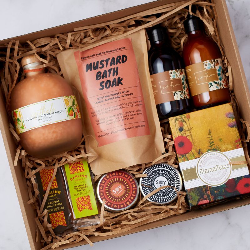 Close-up of a wellness gift box with mustard bath soak, lotions, soy candles, toffees, and Mamamac's shortbread.