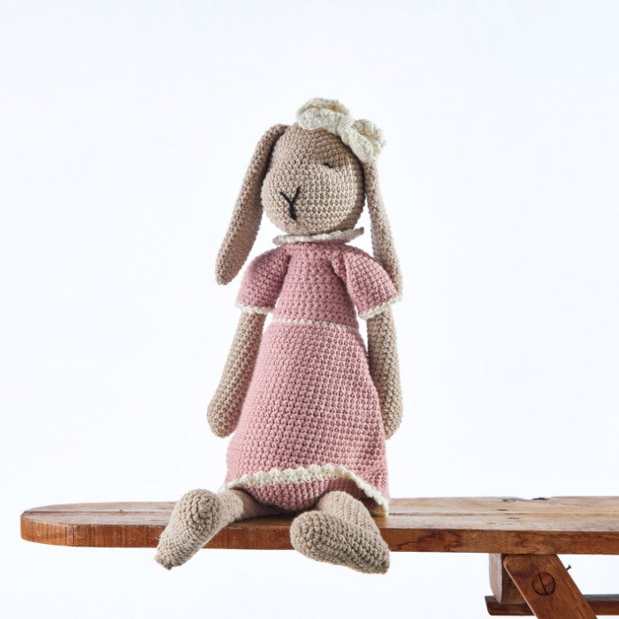 Rosie Rabbit Handmade Doll, a charming crochet rabbit in a pink dress and cream bow, sitting on a wooden bench.