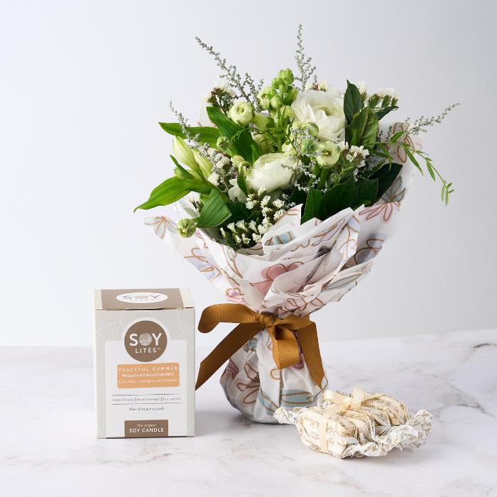 Scent of Summer Gift Set featuring a bouquet of white blooms wrapped in floral paper, a SoyLites Peaceful Summer candle, and Sally Williams nougat on a white surface.