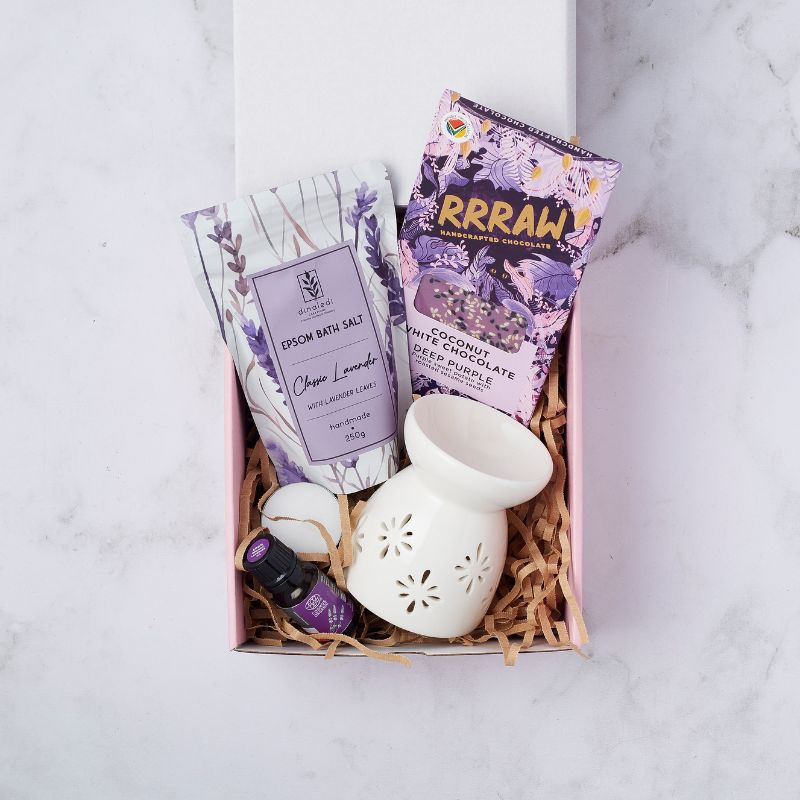 Lavender-themed spa gift box containing Epsom bath salts with lavender leaves, a bottle of lavender essential oil, a white ceramic diffuser, a soft-glow candle, and a handcrafted Rrraw coconut white chocolate bar, arranged on a pink shredded filler.