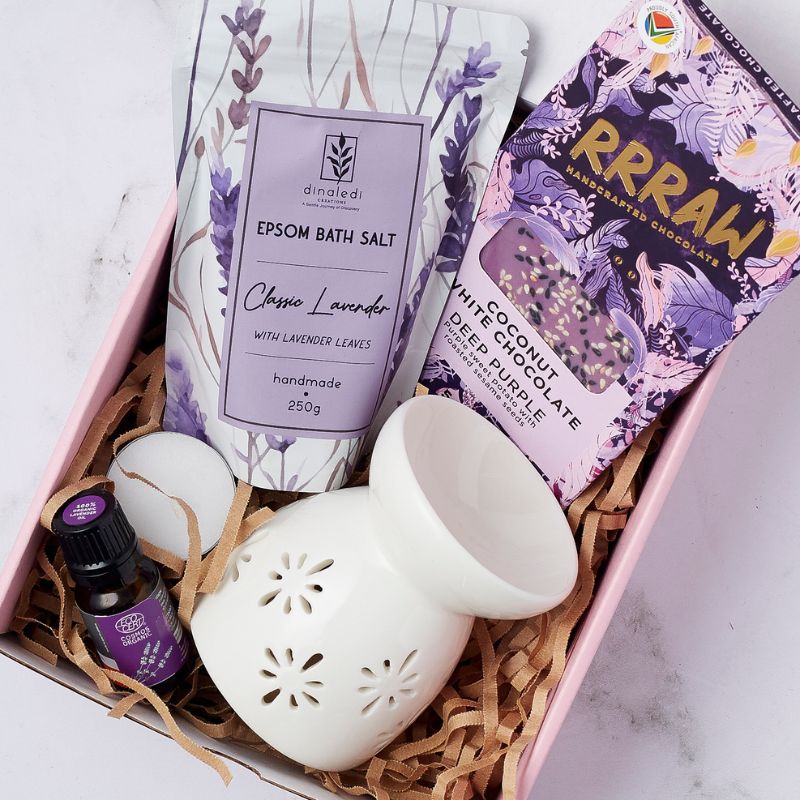 Close-up view of a lavender spa gift set featuring classic lavender Epsom bath salts, lavender essential oil, a white ceramic diffuser, a soft candle, and a handcrafted Rrraw coconut white chocolate bar, elegantly arranged in a pink gift box.