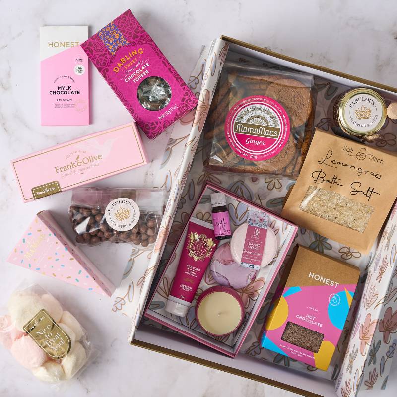 Assorted luxury treats and gifts beautifully arranged in the Sparkle and Sweetness Gift Box, featuring gourmet chocolates, bath salts, ginger biscuits, marshmallows, toffee, and skincare products in a pink and gold themed package.