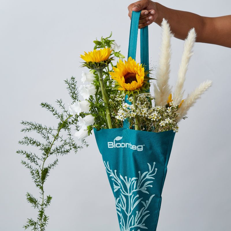 Hand holding a teal Bloombag filled with vibrant sunflowers, white flowers, and greenery.