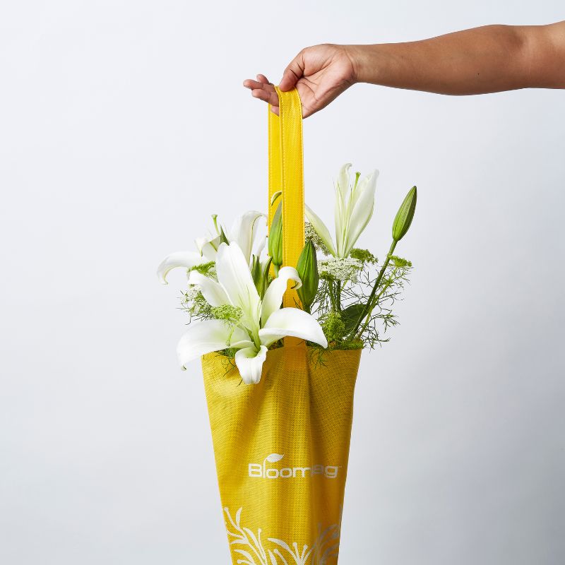 Hand holding a vibrant yellow Bloombag filled with elegant white lilies and lush green foliage.