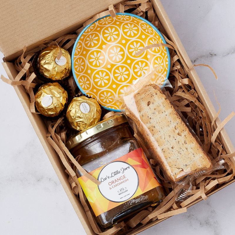 Close-up of gift box contents including Ferrero Rocher chocolates, orange & cardamom jam, yellow ceramic bowl, and biscotti