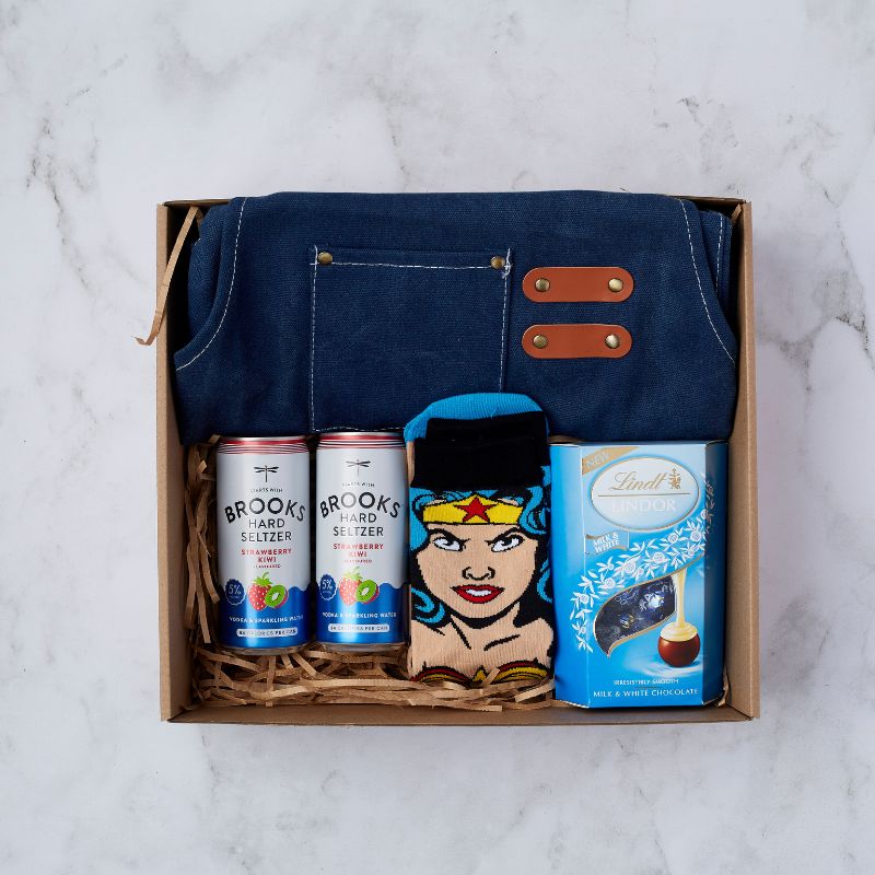 Gift box featuring Brooks Hard Seltzer cans, a blue apron with leather accents, Wonder Woman-themed socks, and Lindt Lindor milk and white chocolate.