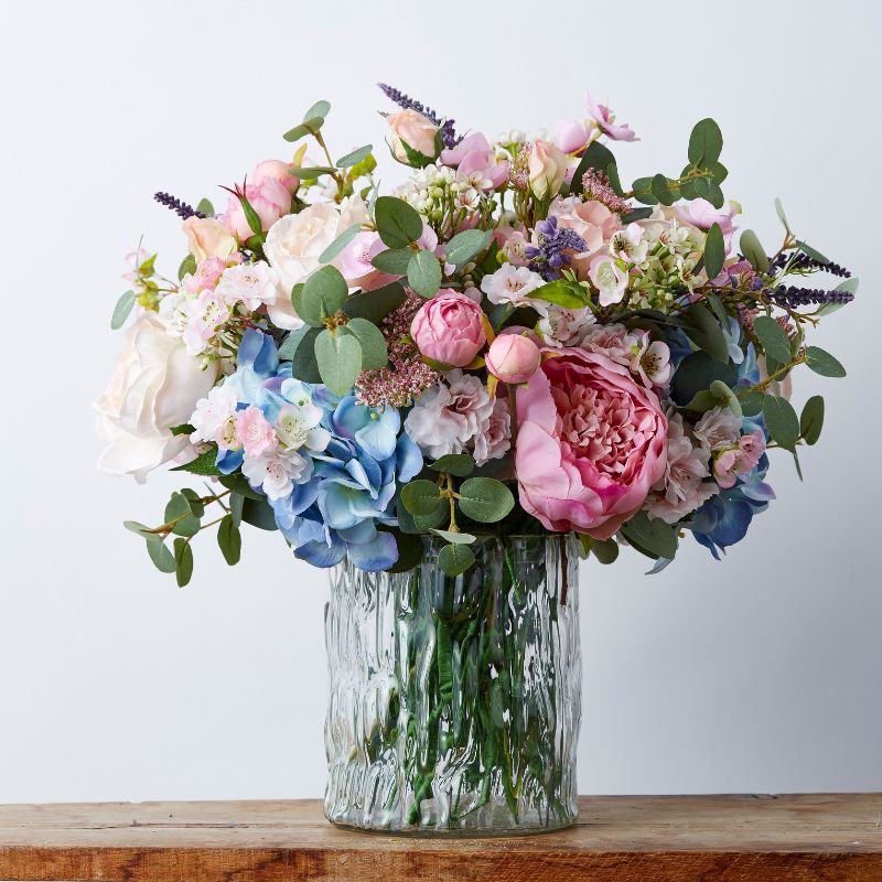 Elegant arrangement of silk flowers featuring soft pink and moody blue blooms, displayed in a textured glass vase, surrounded by lush greenery.